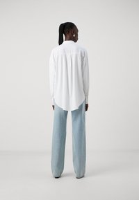 White long-sleeve shirt with a curved hem, paired with light blue wide-leg jeans. Smooth fabric and relaxed fit. Shiny silver earrings.