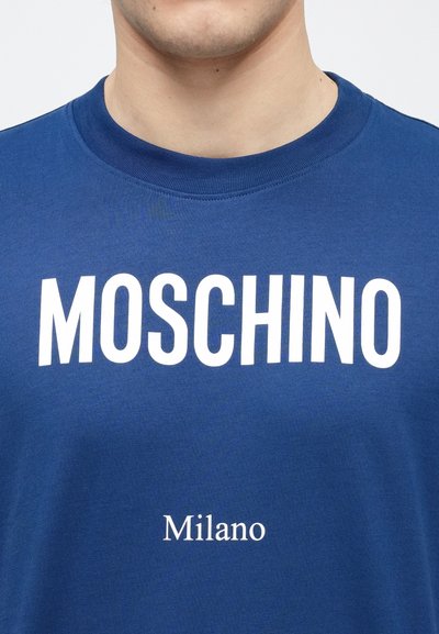 Plava pamučna majica s okruglim izrezom, s velikim bijelim natpisom "MOSCHINO" i manjim natpisom "Milano" ispod, u čistom bezserifnom fontu.
