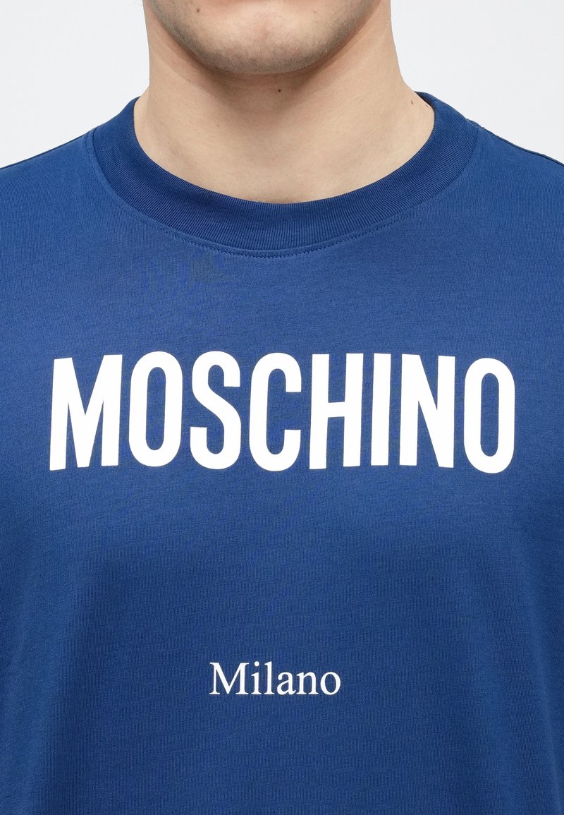 Blauw katoenen T-shirt met een ronde hals, voorzien van grote witte tekst "MOSCHINO" en daaronder kleinere tekst "Milano" in een strak schreefloos lettertype.