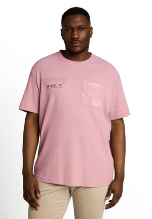 Uomo che indossa una camicia rosa chiaro a maniche corte con taschino sul petto e pantaloni beige, in piedi davanti a uno sfondo bianco.