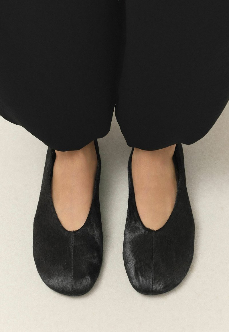 Pieds chaussés de chaussures plates noires, basses, avec une texture lisse légèrement brillante, assorties à un pantalon noir à jambes larges.