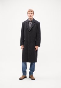 Dunkelgrauer Wollmantel mit kragenloser Halslinie, zwei vorderen Taschen und einem einzigen Knopfverschluss. Über einem grauen Hemd mit Knöpfen und blauen Jeans getragen.