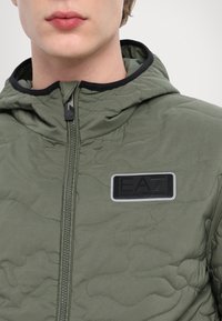Quilted green jacket featuring a black zip closure and hood, textured fabric with a camo pattern, and an embroidered logo patch.
