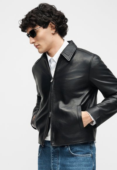 Άνδρας με σγουρά μαλλιά που φοράει μαύρα γυαλιά ηλίου, μαύρο δερμάτινο μπουφάν πάνω από λευκό πουκάμισο και μπλε τζιν, στέκεται με τα χέρια στις τσέπες.