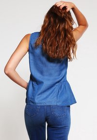 Kvinna med långt vågigt rött hår bär en ärmlös blå denimtopp och blå denimjeans, med handen vilande på huvudet, vänd bort från kameran framför en neutral bakgrund.