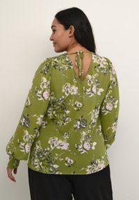 Grøn blomsterbluse med lange, puffede ærmer, v-formet rygudskæring og bindebånd. Den har pink og hvide roser med mørkegrønne detaljer.