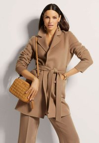 Tan mantel met ceintuur, geknipt revers, lange mouwen en een gestructureerde stof, gecombineerd met een gewatteerde taupe handtas en gouden accessoires.
