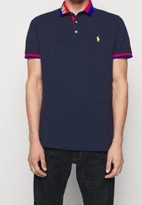 Marinblå polo-shirt med röda och blå krage-accenter, med en liten gul logotyp och tre-knappsknäppning. Kortärmad, tillverkad av bomull.