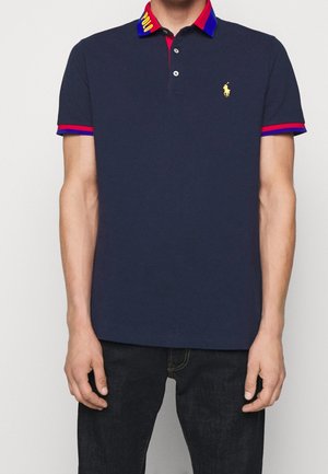 Navy polo shirt met rode en blauwe kraagaccenten, voorzien van een klein geel logo en een drieknopenlaat. Korte mouwen, gemaakt van katoen.