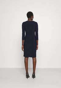 Kvinna med kort hår klädd i en figurnära marinblå knälång klänning och svarta högklackade skor, stående mot en enkel vit bakgrund.