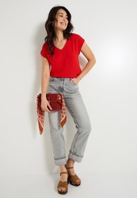 Haut rouge à manches courtes avec un col festonné, jeans délavés aux poignets, sandales à plateforme marron et une pochette rouge avec une écharpe à motifs.