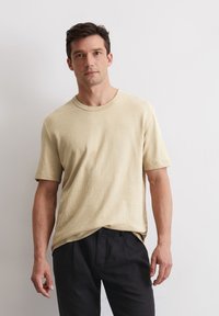 Beige T-Shirt mit kurzen Ärmeln aus einem weichen, leichten Stoff und Rundhalsausschnitt. Kombiniert mit dunklen, lässig geschnittenen Hosen.