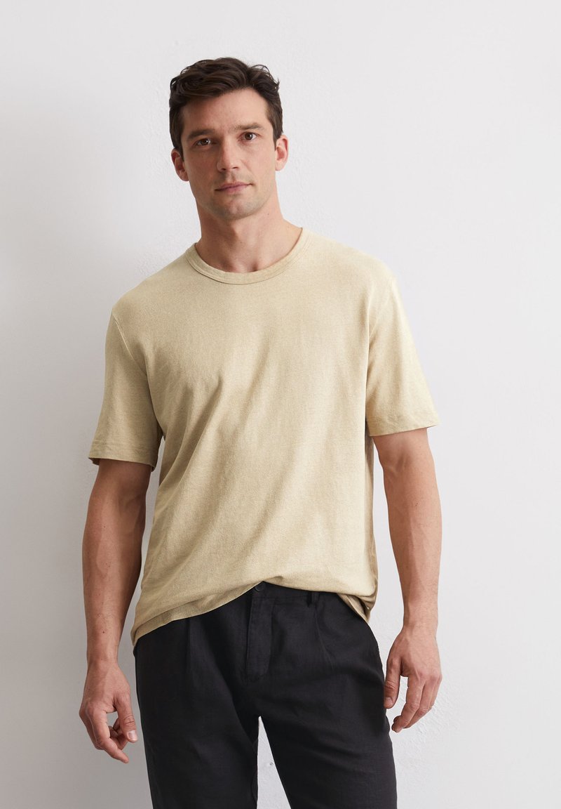 Beige T-Shirt mit kurzen Ärmeln aus einem weichen, leichten Stoff und Rundhalsausschnitt. Kombiniert mit dunklen, lässig geschnittenen Hosen.