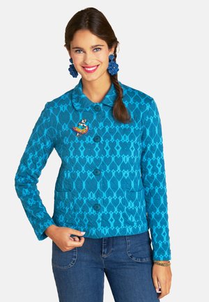 Blue quilted jacket with geometric pattern, front pockets, and round collar. Features a colorful embroidered brooch. Worn with denim.