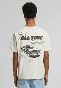 T-shirt en coton beige clair avec un design graphique d'une voiture classique et un texte en gras au dos. Col rond et manches courtes.