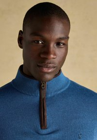 Tuva vīrieša, kurš valkā zilu džemperi ar ceturtdaļas rāvējslēdzēju un brūnu apmali, ar mazu izšūtu logotipu uz krūtīm, neitrāls bēšs fons.