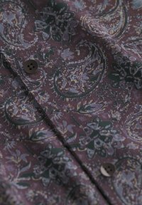Dunkelvioelner Stoff mit Paisley- und Blumenmustern in gedämpftem Blau und Gelb, mit zwei dunkelnen Knöpfen an einem gefalteten Hemdteil.