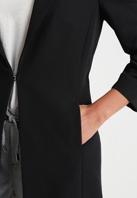 Schwarze Blazer mit einem einzelnen Reißverschluss, langen Ärmeln und Seitentaschen. Der Stoff wirkt glatt und strukturiert mit einem taillierten Design.