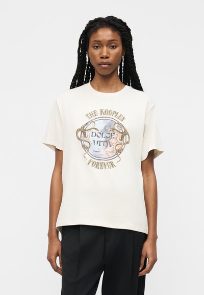 Femme aux longues tresses portant un t-shirt blanc avec un texte graphique doré et bleu "The Kooples Dolce Vita Forever" et un pantalon noir, debout devant un fond blanc.