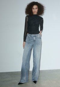 Schwarzes Langarmoberteil mit Netz- und Punktemuster, kombiniert mit weit geschnittenen hellblauen Jeans und spitzen schwarzen High Heels. Schlichter Hintergrund.