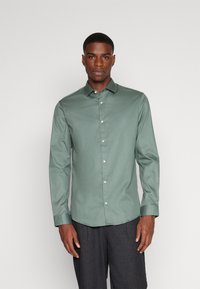 Chemise verte à manches longues, boutonnée, en tissu lisse, avec un col classique, des boutons blancs et une coupe ajustée.