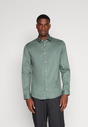 Camisa verde de manga larga con botones, hecha de una tela suave, que presenta un cuello clásico, botones blancos y un corte entallado.