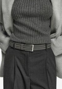 Persoon in een grijze ribbeltrui gestoken in donkere geplooide broek met een gevlochten tekstuurbelt en een lichtgrijze jas, met de handen in de zakken.