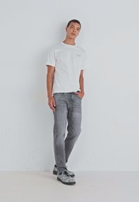 Γκρι τζιν, λευκό μπλούζα με στρογγυλή λαιμόκοψη και γκρι παπούτσια με μαύρες κορδόνες. Το μοντέλο στέκεται ενάντια σε ένα απλό λευκό φόντο, με τα χέρια στις τσέπες.