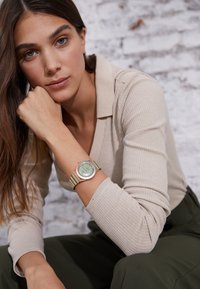 Montre verte et dorée avec un cadran rond et un bracelet rayé, portée au poignet. Le sujet a les cheveux longs et porte un haut de couleur neutre devant un fond.
