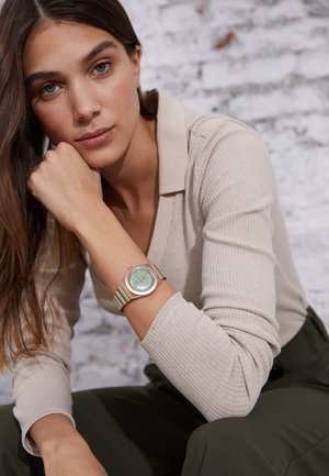 Green and gold watch with a round face and striped band, worn on a wrist. Subject has long hair and is in a neutral-colored top against a backdrop.