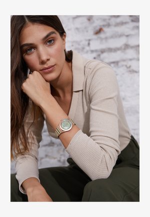 Green and gold watch with a round face and striped band, worn on a wrist. Subject has long hair and is in a neutral-colored top against a backdrop.