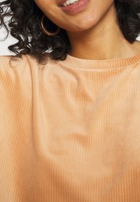 Vrouw glimlachend in een perzikkleurig geribbeld topje en gouden ringoorbellen, met krullend donker haar, zichtbaar tegen een witte achtergrond.