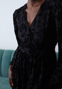 Czarna sukienka kopertowa w kwiaty z głębokim dekoltem w serek, długimi przezroczystymi rękawami i wiązaniem w talii. Materiał o fakturze z subtelnym kwiatowym wzorem.