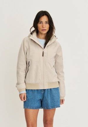 Vrouw draagt een beige jas met capuchon over een lichtblauwe trui en spijkershorts, staand tegen een effen lichte achtergrond.