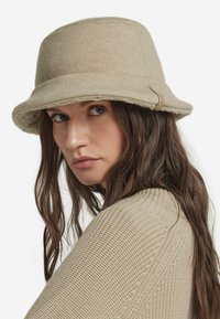 Beige filthatt med mjuk kant, har en texturerad yta och en sidetikett. Modellen har långt, vågigt brunt hår och bär en stickad tröja.