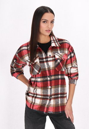 Red, brown, and white plaid flannel shirt with two front pockets, button closure, and rolled-up sleeves, worn over a black shirt.
