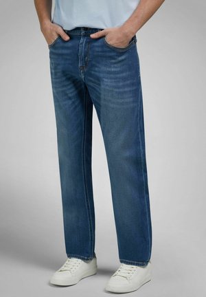 Mann trägt gerade geschnittene blaue Jeans und weiße Turnschuhe, die Hände in den Taschen, vor grauem Hintergrund.