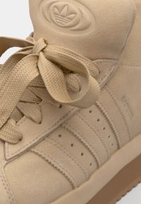Sneaker alta in camoscio beige con pannelli testurizzati, lacci piatti e un logo sulla linguetta. Suola in gomma con un colore a contrasto sul fondo.