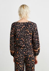 Blouse noire à motif floral avec des manches longues et des poignets élastiques, offrant une coupe décontractée. Les détails incluent un ourlet rayé contrasté.