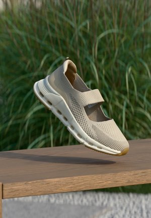 Hellbeiger atmungsaktiver Strick-Sneaker mit Klettverschluss, dicker weißer Sohle, schwebt über einer Holzoberfläche mit grünem, verschwommenem Pflanzenhintergrund.