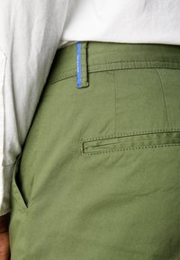 Zielone oliwkowe spodnie wykonane z tkaniny mieszanej bawełny, z niebieską metką na pasie i tylną kieszenią z eleganckimi detalami przeszycia.
