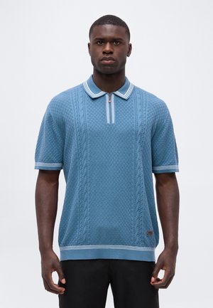 Homme portant un polo bleu texturé à manches courtes avec un col rayé blanc et une fermeture éclair, debout devant un fond uni.