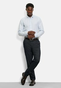 Hellblau gestreiftes Langarmhemd, dunkle gemusterte Hose, brauner Ledergürtel und glänzende braune Schuhe. Das Modell steht lässig mit verschränkten Armen.