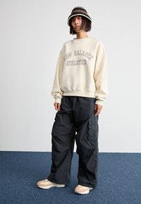 Beige Sweatshirt mit dem Aufdruck "New Balance Athletics" in Braun, kombiniert mit schwarzen Cargo-Hosen. Beigefarbene und weiße Sneakers mit strukturierten Sohlen.