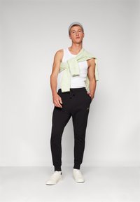 Schwarze Jogginghosen mit elastischen Bündchen, kombiniert mit einem weißen Tanktop. Ein hellgrüner Sweatshirt ist über die Schultern gelegt. Weiße Sneaker und eine graue Mütze vervollständigen den Look.