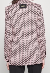 Blazer de patrón rosa y negro con formas geométricas, solapas en forma de muesca y una etiqueta en la parte posterior. Mangas largas con detalles de botones.