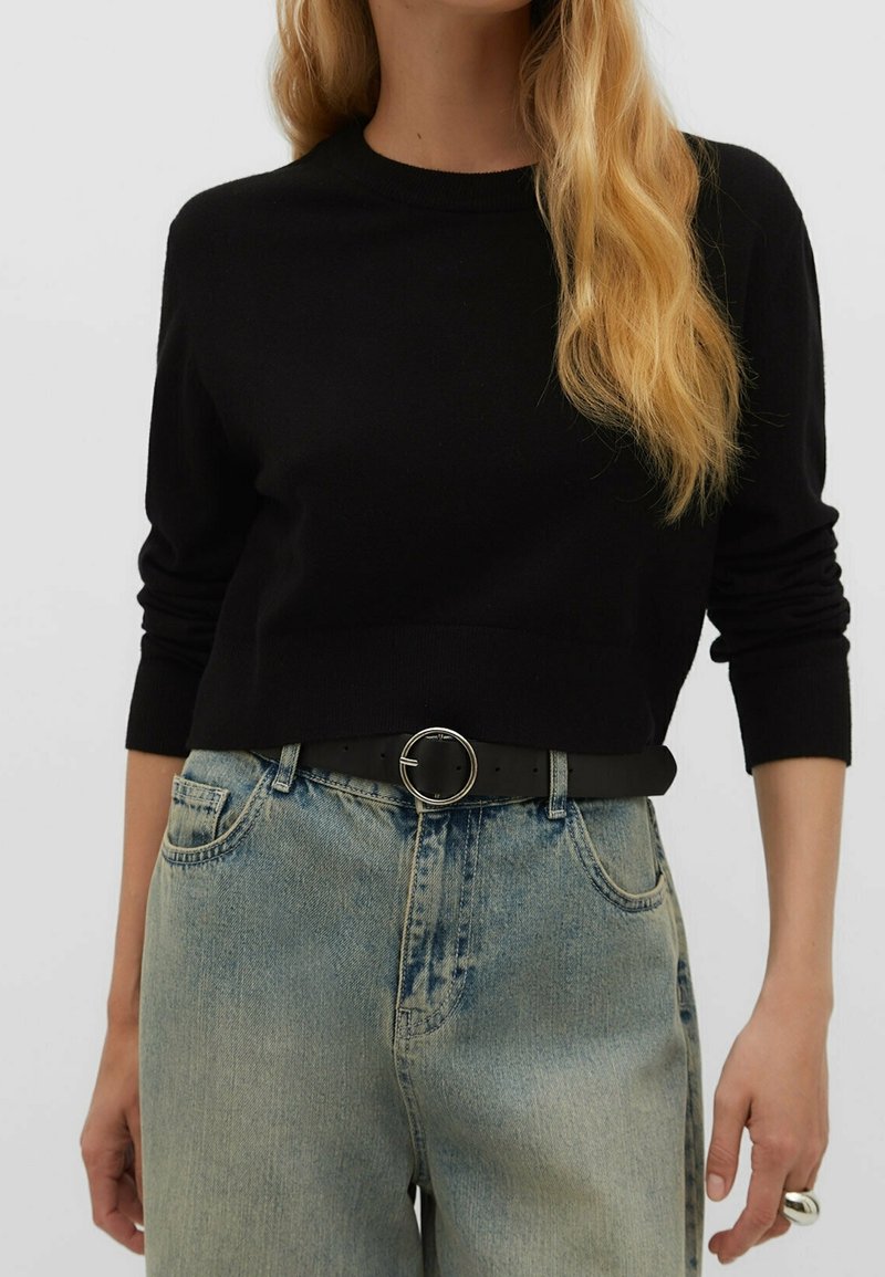 Femme portant un pull court noir à manches longues, une ceinture noire avec une boucle ronde en argent, et un jean taille haute bleu clair.