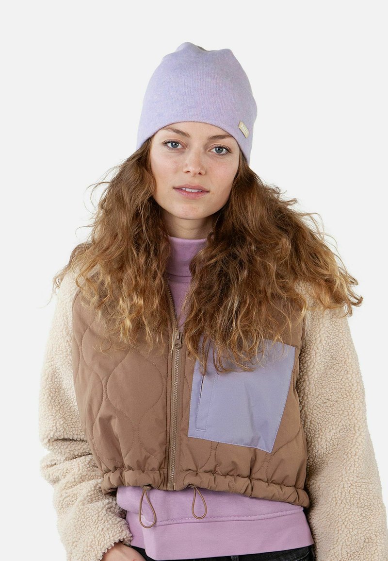 Brown quilted vest with a lilac accent pocket, paired with a light purple beanie and a pastel pink sweatshirt, featuring curly hair.