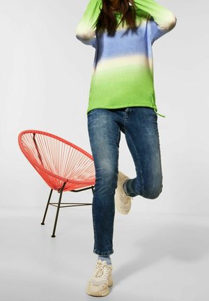Green and blue gradient pullover, lightweight knit, paired with dark denim jeans and chunky white sneakers. Red accent chair in the background.