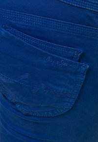 Jeans in denim blu con una tasca posteriore decorata da cuciture e un sottile logo del marchio. Il tessuto ha una consistenza liscia e un design aderente.